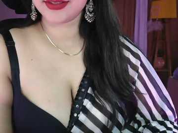A video of a woman with fair skin and long black hair, wearing a black bra and a striped, open-front shirt. She has red lipstick and dangling earrings. The background is a softly lit room with a lamp.