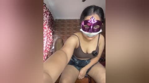 A video of a young woman with medium skin tone, wearing a purple mask and denim shorts, sitting on a bed in a bedroom with red curtains and a brick wall.