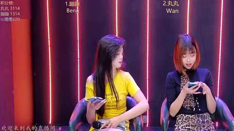 Video of two young women, one with long black hair and a yellow T-shirt, the other with a bob haircut and a leopard-print dress, seated in chairs, watching a screen, in a dimly lit room with neon lights.