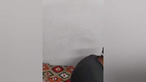 Video of a person in a grey shirt, with their head slightly tilted to the right, standing near a plain white wall with a small, faint smudge. Background shows a patterned blanket with red, orange, and white geometric shapes.