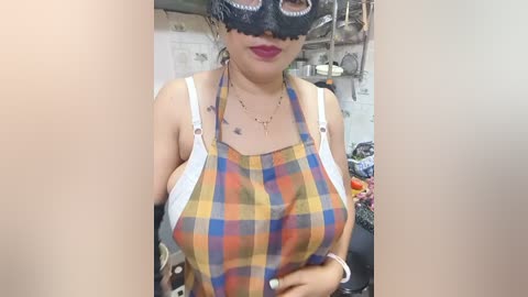 Video of a woman with medium skin tone, wearing a black mask, plaid apron, and red lipstick, standing in a cluttered kitchen.