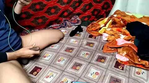 Video of a person wearing a blue shirt, seated on a bed with a patterned quilt, surrounded by colorful clothing and an electronic device.