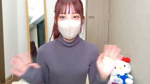 A video of an Asian woman with long brown hair and bangs, wearing a gray turtleneck, white face mask, and red gloves. She stands in a modern room with light-colored walls and a Hello Kitty plush toy on the right.