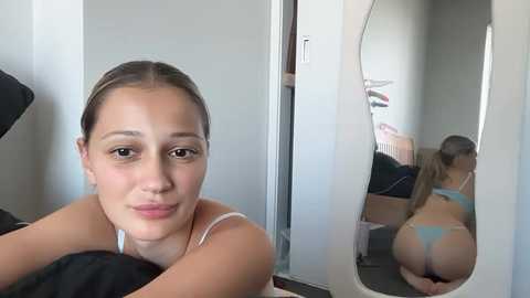 A video of a young woman with light skin, brown hair tied back, wearing a white bra and blue thong, smiling into a mirror. She is seen from the front and rear, showing her bare buttocks.