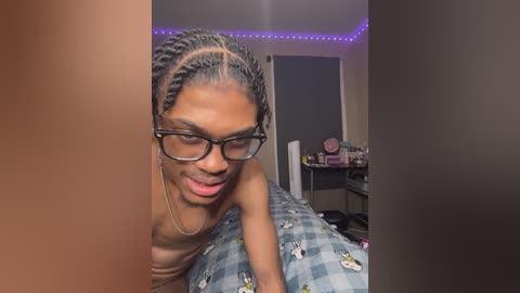 Video of a topless, smiling Black transgender woman with braided hair, wearing glasses, lying on a bed with a checkered blue and white duvet. Background shows a cluttered room with a desk, purple LED strip lights, and a closed door.