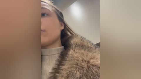 A close-up video of a woman's face and shoulders, partially obscured by a blurred, beige-colored object. She wears a beige turtleneck sweater and has light brown hair. The background is a neutral-toned room with a ceiling light.