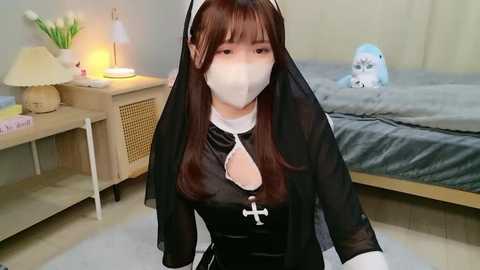 Video of an Asian woman in a black habit, face mask, and veil, kneeling in a bedroom with a bed, nightstand, and lamp.