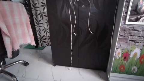 Video of a black suitcase with a white cord hanging from its handle, placed against a wall adorned with a colorful floral wallpaper. A pink and white striped blanket covers a chair in the background.