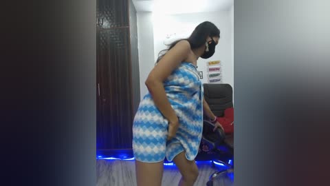 Video of a curvy woman with medium skin tone, wearing a blue and white striped towel, standing in a dimly lit room with a black chair and colorful wall art.