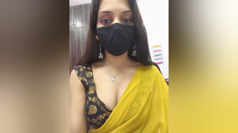 Video of a South Asian woman with long dark hair, wearing a yellow saree, black mask, and a black blouse with a floral pattern. She has a silver necklace and earrings. Background shows a blurred indoor setting.