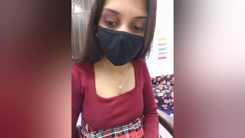 Video of a young South Asian woman with medium brown skin, wearing a black face mask, red lipstick, and a maroon top with a plaid skirt. Background shows a room with a floral-patterned blanket and a white wall.