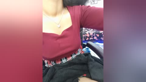 Video of a woman with medium skin tone wearing a maroon top with a plunging neckline, black skirt, and a floral-patterned blanket. She's seated in an office chair, partially obscured by a red and purple gradient overlay.