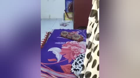 A video showing a simple room with a purple bedspread featuring large pink flowers, a polka-dotted curtain, and a small chair.