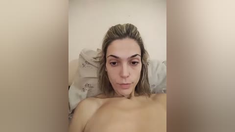 Video of a nude woman with light skin, shoulder-length blonde hair, and small breasts lying on a pillow in a dimly lit room with a beige wall.