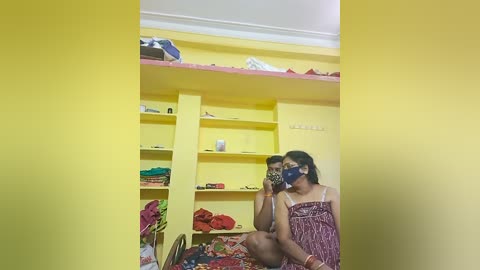 Video of a brightly lit, yellow-painted room with two people in casual attire; one is wearing a face mask. Shelves hold clothes and personal items.