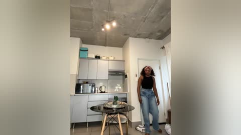 Video of a modern, minimalist kitchen with exposed concrete ceiling, light gray cabinets, and a woman in a black top and light-wash jeans, standing near a small glass table.