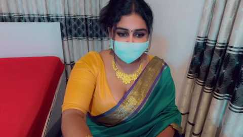 A video of a South Asian woman with medium skin tone and dark hair, wearing a yellow blouse, green sari with gold border, and a turquoise mask, sitting on a red bed with patterned curtains in the background.