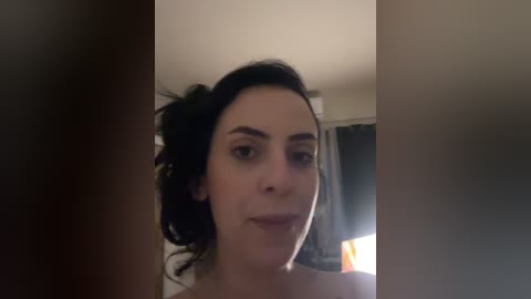 Video of a fair-skinned woman with dark hair tied back, smiling slightly. She's indoors with dim lighting, partially visible curtains, and a slightly blurred background.