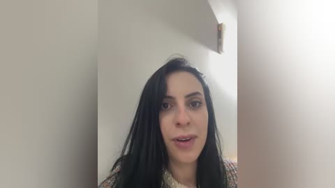 Media: Video of a young woman with long black hair, wearing a floral-patterned top, standing in a white-walled room with a corner visible. She has a neutral expression.