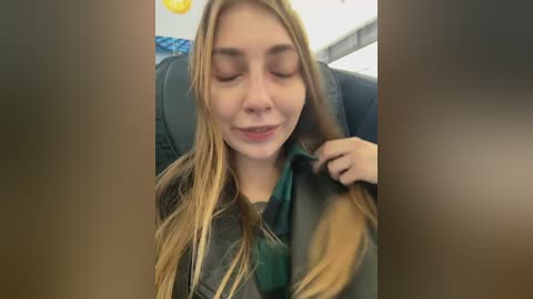 Video of a young Caucasian woman with long blonde hair, closed eyes, and a slight smile, sitting in a dark car with a green scarf around her neck.