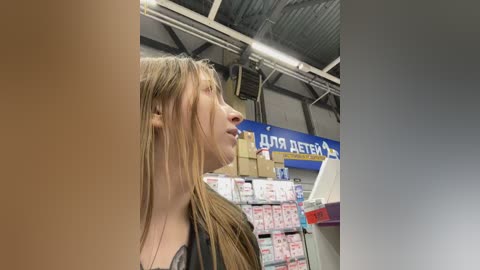 Video of a young woman with long blonde hair, light skin, and a black top, standing in a cluttered, industrial storage room with shelves full of boxes and a blue sign reading \"DOSYAL DETERGENT.\