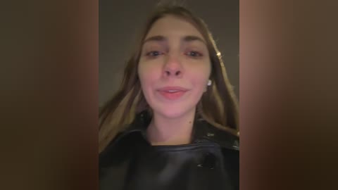 A video of a young woman with long brown hair, light skin, and a black leather jacket, smiling slightly, with a blurred background.