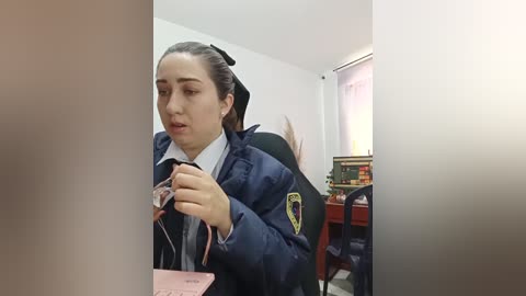 Video of a young woman with dark hair in a ponytail, wearing a navy blue school uniform, seated at a desk, holding a pink item. Background shows a classroom with books and a window.