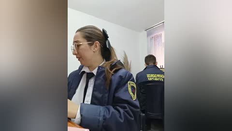 Video of a female police officer with glasses, dark hair in a ponytail, wearing a navy blue uniform, white shirt, and black tie, standing in a room with another officer in the background.