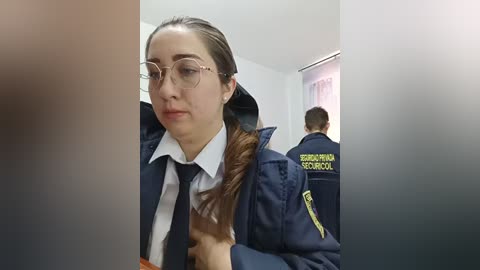 Video of a young woman with glasses in a navy police uniform with a black tie and a hood, standing in a hallway with another officer. Background includes a partially visible second officer.