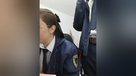 Video of an Asian woman with glasses, dark hair tied back, wearing a navy-blue police uniform with a yellow badge, seated at a desk in a dimly lit room.