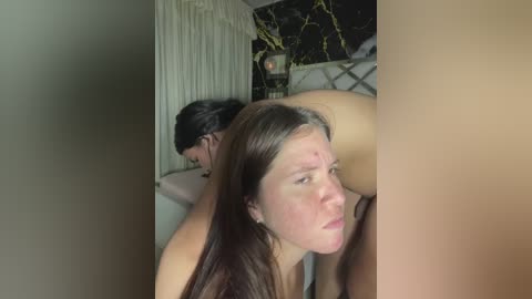 Video of a woman with long brown hair, fair skin, and freckles, lying on her back with a man behind her. The background features a white curtain and a decorative plant.