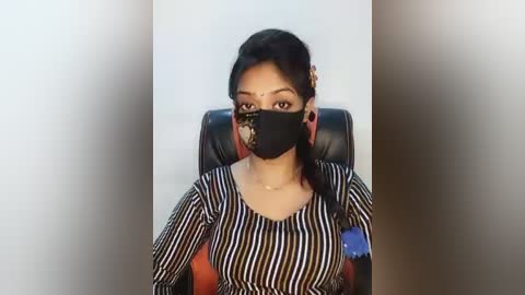 Video of a woman with medium-dark skin and black hair in a side braid, wearing a black face mask, black-and-white striped top, and sitting in a black chair.