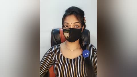 Video of a woman wearing a black face mask, black-and-white striped long-sleeve top, and brown leather crossbody bag. She has medium brown skin and dark hair, sitting in a black chair against a light gray wall.