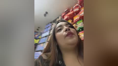 Video of a young Hispanic woman with light brown skin, straight brown hair, and full lips, standing in a brightly lit supermarket aisle filled with colorful packaged products, including cereals and snacks.