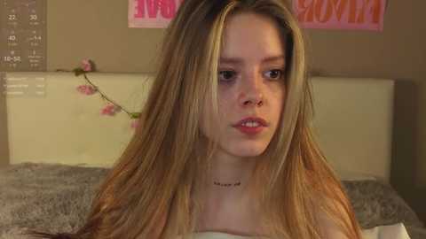 Video of a young Caucasian woman with long, blonde hair, fair skin, and light makeup, wearing a black choker. She sits on a bed with grey bedding in a softly lit bedroom.