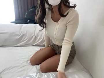 Video of an Asian woman with long black hair, wearing a white face mask, beige sweater, and plaid skirt, kneeling on a white bed with a black chair in the background.