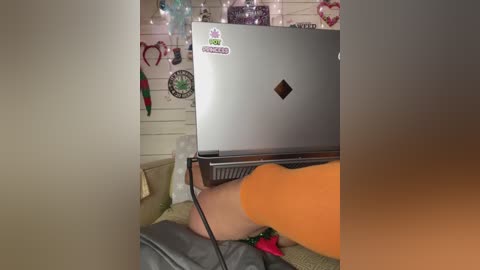 Video of a person in an orange shirt sitting on a grey couch, legs propped on a silver laptop, with a cozy, cluttered background of fairy lights, wall decor, and a white shag rug.
