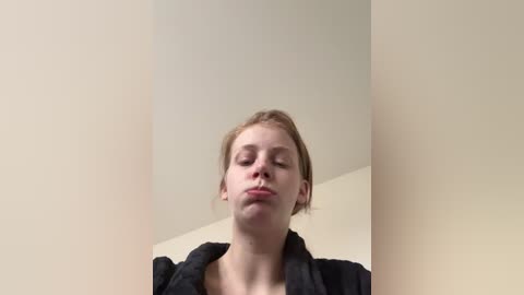 A video of a young woman with fair skin and blonde hair, wearing a black robe, making a pouty face. The background is a plain, light beige wall.