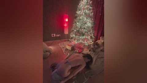 A video shows a woman in a dark room, kneeling near a lit Christmas tree, wearing only a blue wristband, with a faint red light casting a warm glow.