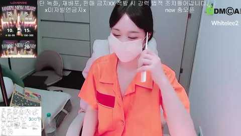 A video of an Asian woman in a surgical mask, orange scrubs, and headphones, sitting in a dimly lit room, with a TV displaying Korean news in the background.