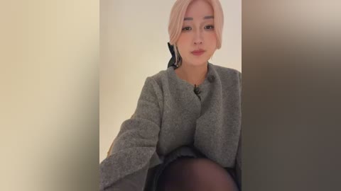 Media: Video of a young Asian woman with fair skin, platinum blonde hair, and light makeup. She wears a grey wool coat over a black top and dark tights, seated in a dimly lit, beige-walled room.