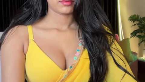 Video of a South Asian woman with long black hair, wearing a yellow blouse with a low neckline, showcasing medium-sized breasts, in an indoor setting with a green plant and a dark curtain in the background.