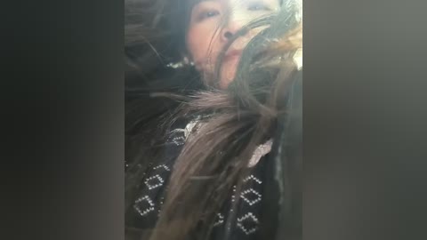 Media: Video of a young woman with long, messy brown hair partially obscuring her face, wearing a black jacket with white geometric patterns. She has fair skin and a serious expression. The background is dark and blurry.