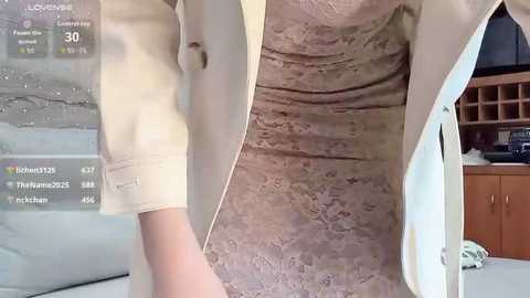 Video of a person wearing beige pants and a white coat, standing in a room with a textured beige wall, wooden shelves, and a TV.