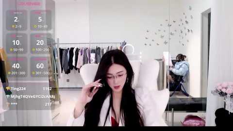 Video of an Asian woman with long black hair, glasses, and a white jacket, sitting in a modern, white-themed clothing store. She's adjusting her hair. Background shows hanging clothes and a mannequin.