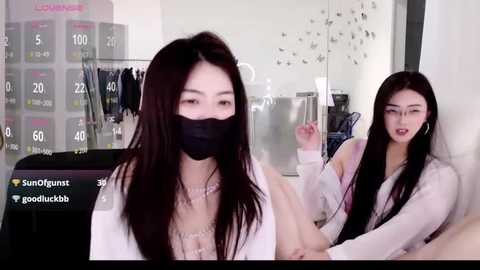 Video of an Asian woman with long black hair, wearing a black mask, white top, and white cardigan, seated in a modern room with a stainless steel fridge and a display rack of clothes in the background.