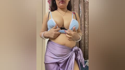 Video of a plus-sized woman with medium skin tone, wearing a blue lace bra and purple sarong, lifting her top to reveal ample breasts, in a dimly lit room with beige walls.