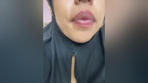 Media: Video of a person's lower face and upper chest, wearing a black hijab and pink lipstick, against a blurred background.
