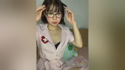 Video of an Asian woman with short, dark hair, wearing glasses, a pink nurse outfit, and adjusting her glasses. Background features a bed with plush toys.