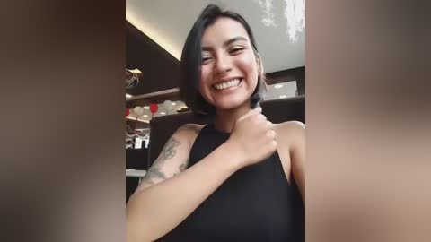 Video of a smiling, dark-haired woman with tattoos on her arm, wearing a black sleeveless top, seated in a dimly lit restaurant with wooden tables and chairs.